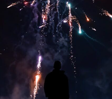 A person silhouetted against a colorful night sky with fireworks bursting overhead.